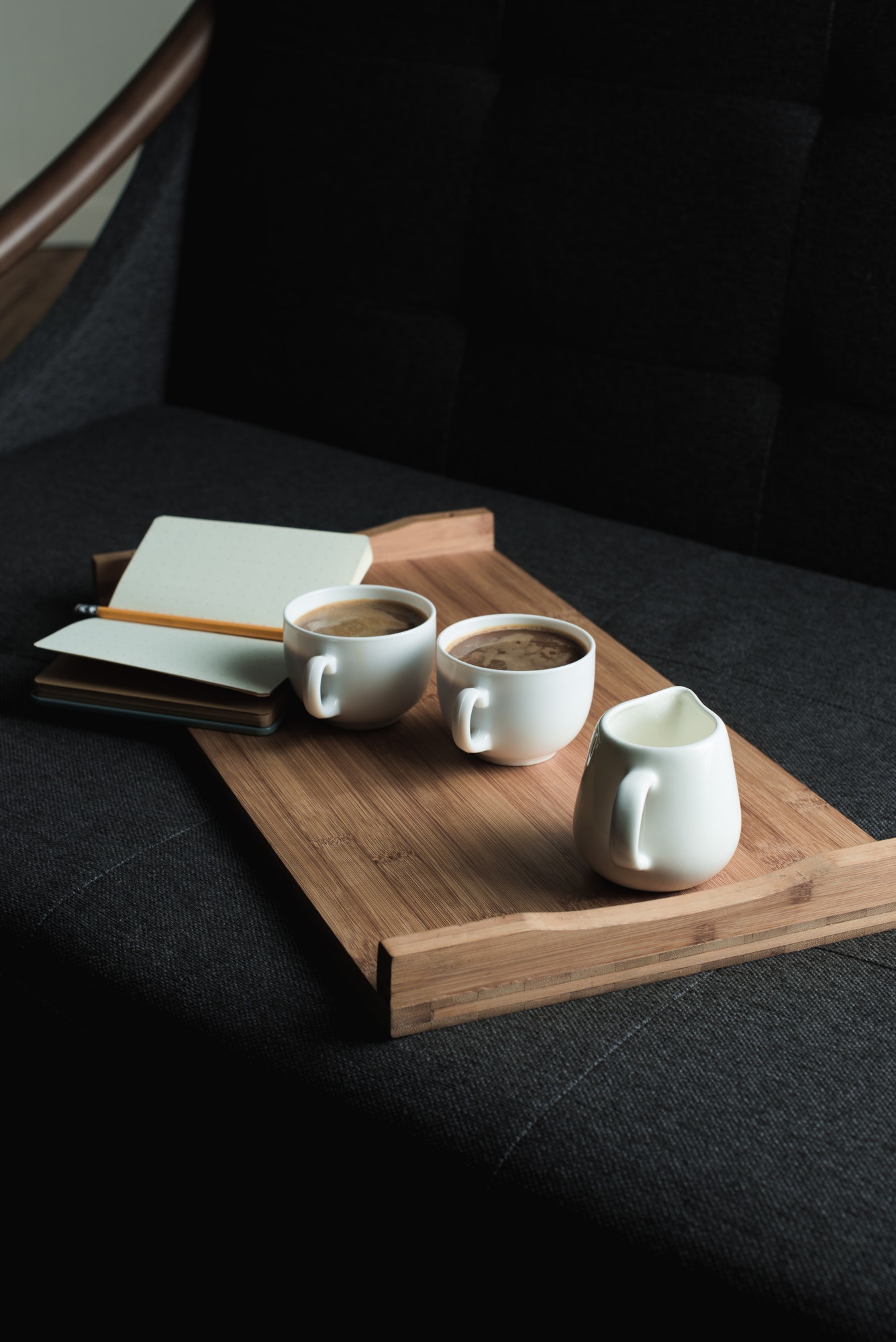 two-cups-of-coffee-jug-of-milk-and-notebook-with-pencil-on-wooden-tray-on-sofa-1